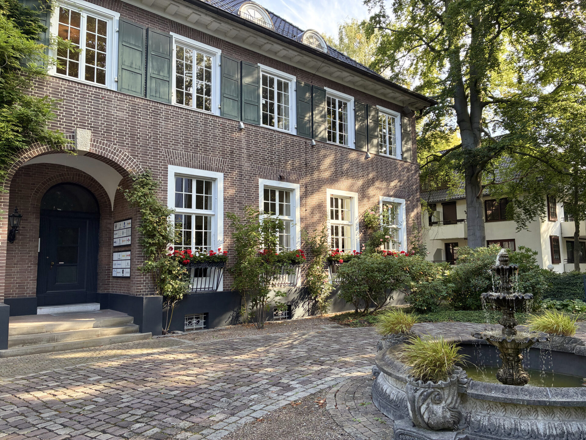 Historisches Gebäude mit Backsteinfassade und grünen Fensterläden, umgeben von einem gepflegten Garten und einem dekorativen Brunnen. Die Fassade ist mit roten Blumen geschmückt, während die Sonne durch die Bäume scheint und eine einladende Atmosphäre schafft.