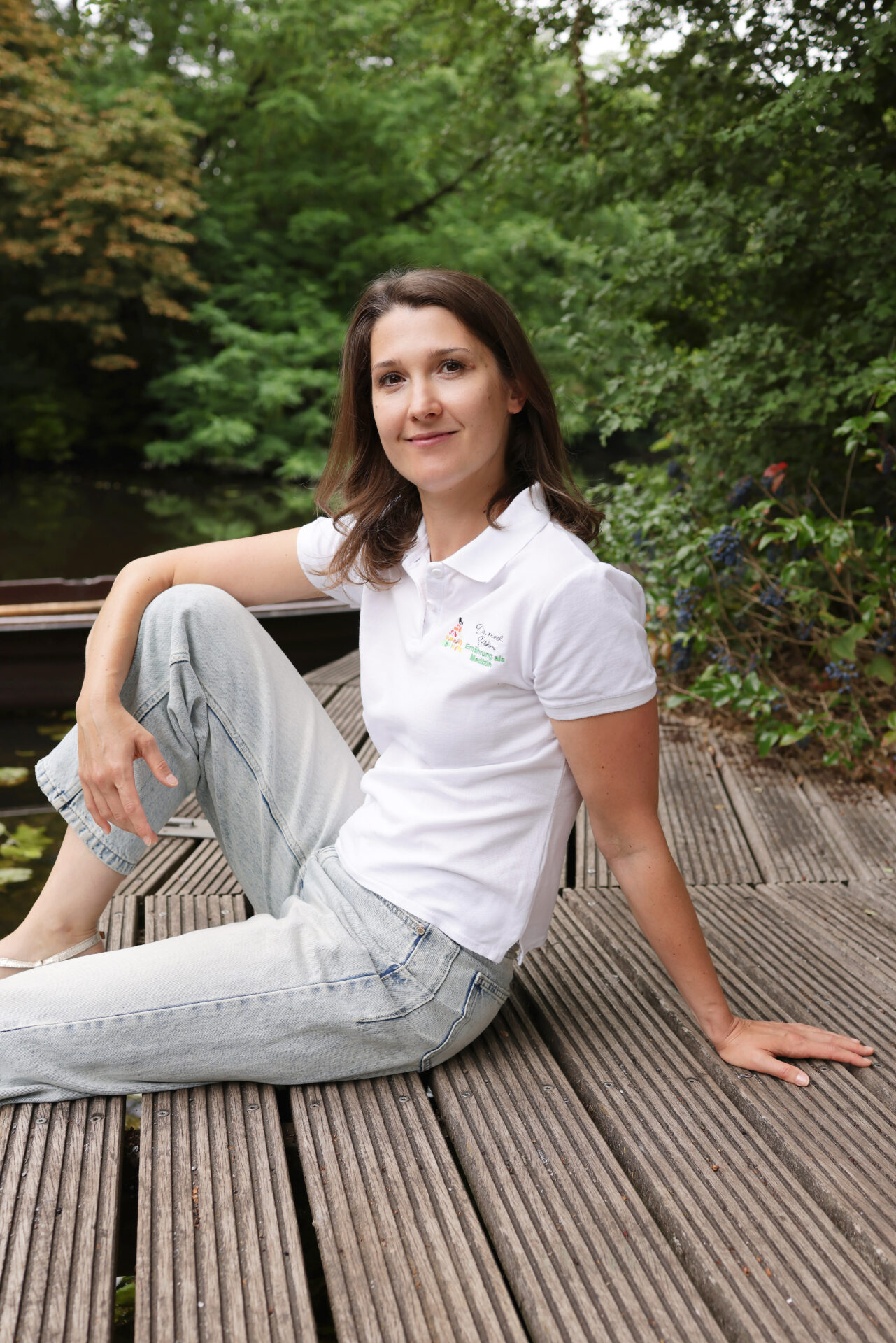 Frau sitzt entspannt auf einem Holzsteg am Wasser, trägt ein weißes Polo-Shirt und helle Jeans. Im Hintergrund ist üppiges Grün zu sehen, das eine natürliche und ruhige Atmosphäre schafft.