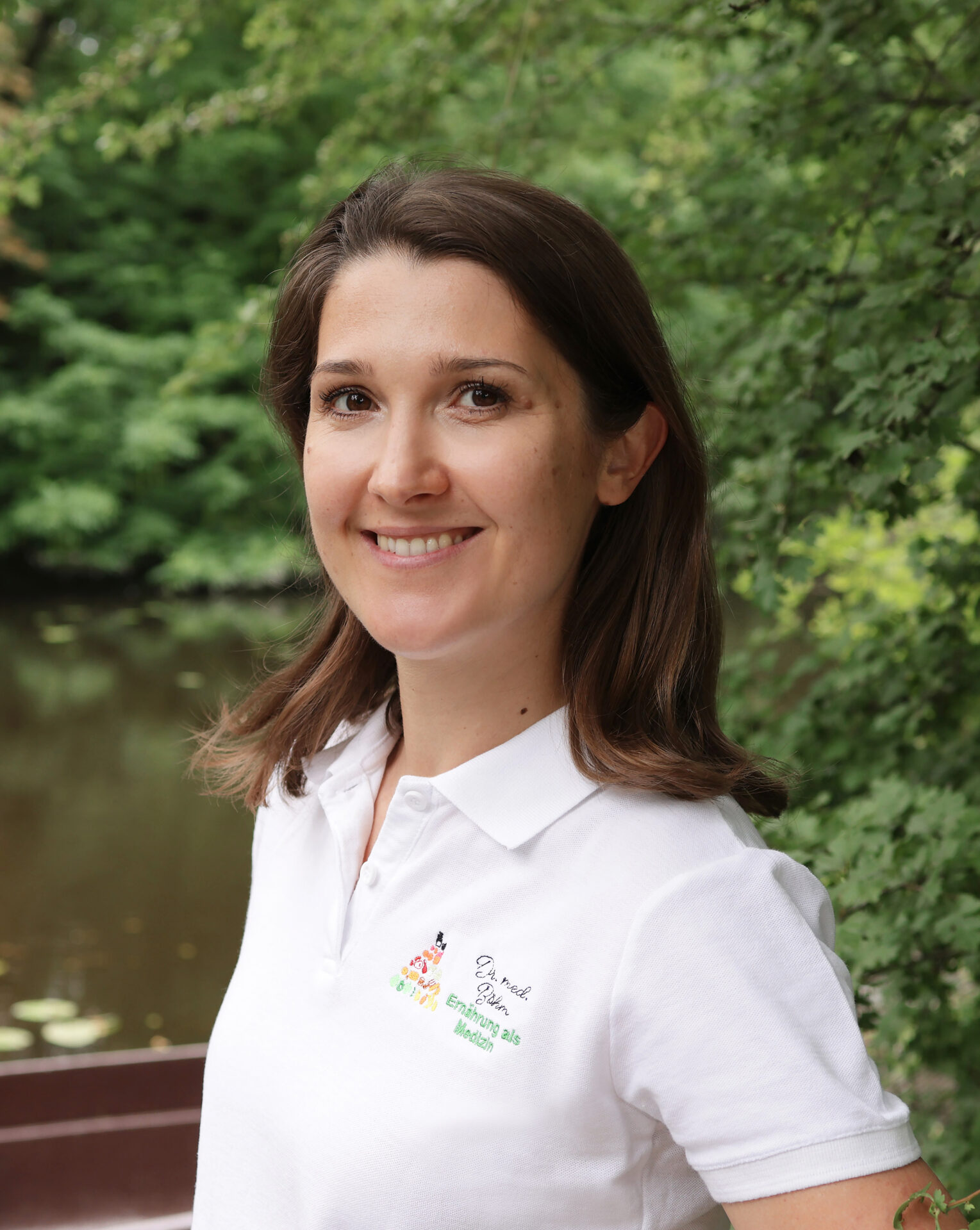 Lächelnde Frau in weißem Poloshirt, die draußen in der Nähe eines ruhigen Teichs steht, umgeben von grüner Vegetation, und eine Botschaft über gesunde Ernährung vermittelt.